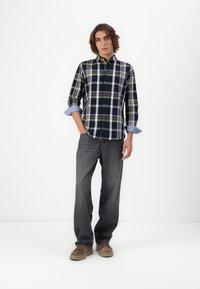 Hemd mit Knopfleiste in Marineblau und Weiß kariert mit grünen Akzenten, hochgerollten Ärmeln, kombiniert mit dunkelgrauen Jeans und braunen Wildlederschuhen.