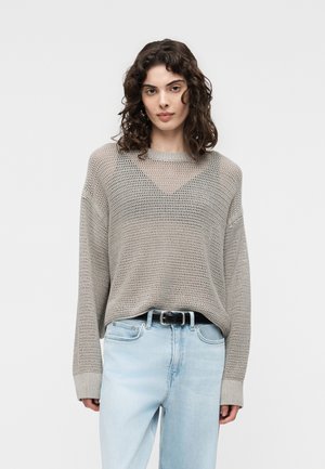 Kvinde med krøllet hår iført en løs, lysegrå striktrøje over en sort top, stoppet ned i lyseblå jeans med et sort bælte.