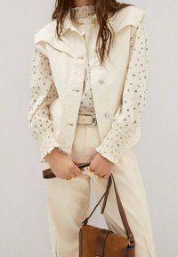 Krémová denimová vesta se strukturovaným límcem přes světle květinově vzorovanou blůzu. Modelka drží hnědou crossbody kabelku a má na sobě béžové kalhoty.