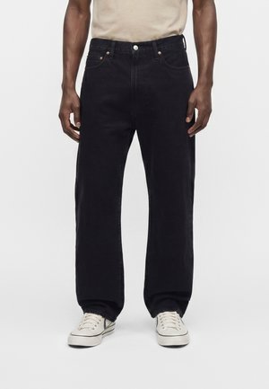 Homme portant un jean noir ample, des baskets blanches et une chemise beige, debout devant un fond blanc uni.