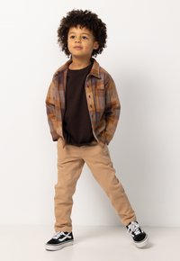 Bruin geruit overhemd over een bruine t-shirt, gecombineerd met beige broek en zwarte sneakers met witte accenten. Kind poseert vol zelfvertrouwen.