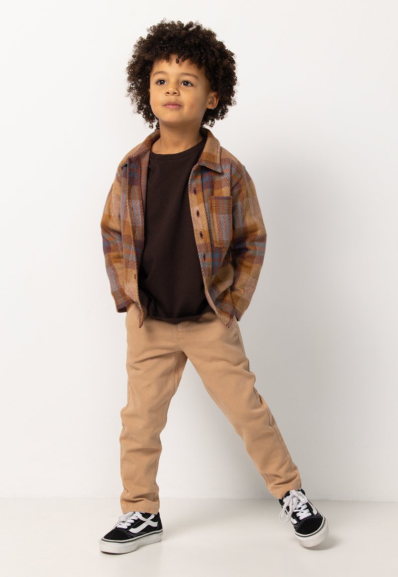 Bruin geruit overhemd over een bruine t-shirt, gecombineerd met beige broek en zwarte sneakers met witte accenten. Kind poseert vol zelfvertrouwen.