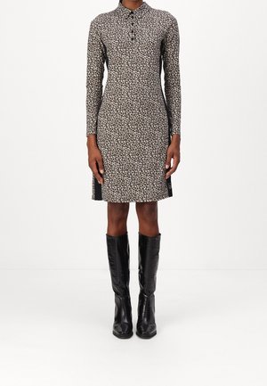 Mujer que lleva un vestido de manga larga con estampado de leopardo y cuello abotonado, combinado con botas de cuero negras hasta la rodilla, de pie sobre un suelo blanco.
