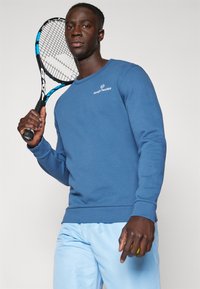 Sudadera azul de mangas largas y puños de canalé, con un pequeño logotipo blanco en el pecho. El modelo sostiene una raqueta de tenis y una pelota.