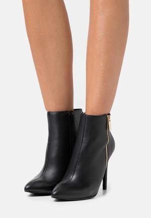 Bottes noires en cuir jusqu'à la cheville avec un bout pointu et un talon aiguille haut. Présente un accent de fermeture éclair doré sur le côté extérieur. Texture lisse.