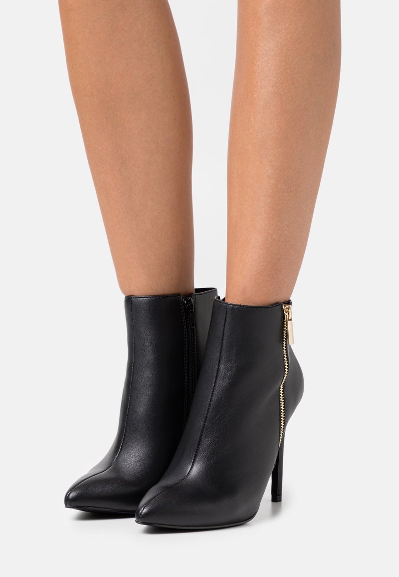 Botines de cuero negro hasta el tobillo con punta afilada y tacón de aguja alto. Presentan un detalle de cremallera dorada en el lado exterior. Textura suave.