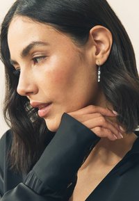 Tumejalgadega naine kannab riputavate kristalleheteid ja musta särki, õrnalt kaela puudutades ja allapoole vaadates.