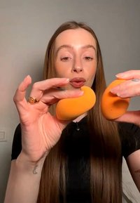 Femme aux longs cheveux tenant et comparant deux éponges à maquillage orange devant un fond neutre.