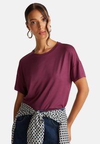 Tričko s krátkým rukávem a kulatým výstřihem z tmavého švestkového materiálu, nošeno s bílo-námořnickou vzorovanou šerpou kolem pasu. Hladká textura, pohodlný střih.