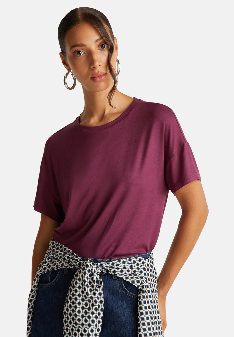 Tričko s krátkým rukávem a kulatým výstřihem z tmavého švestkového materiálu, nošeno s bílo-námořnickou vzorovanou šerpou kolem pasu. Hladká textura, pohodlný střih.