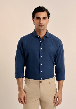 Camisa de botones azul marino hecha de tela texturizada, con cuello, mangas enrolladas y un pequeño logo en el pecho. Pantalones beige.