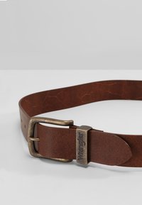 Brauner Ledergürtel mit einer gewerblichen Optik, ausgestattet mit einer rechteckigen Metallschnalle, die mit dem Markennamen "Wrangler" graviert ist.