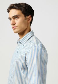 Camicia leggera in cotone con strisce verticali blu e crema, colletto a bottoni e una sottile texture del tessuto.