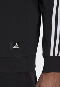 Felpa nera Adidas con tre strisce bianche lungo le maniche, polsini a costine e un'etichetta nera sul margine inferiore.