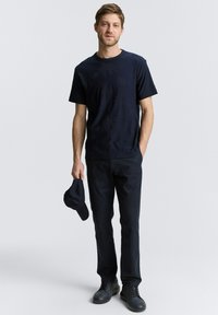 Donkerblauwe T-shirt met subtiel palmbomenpatroon, gecombineerd met donkere broek en zwarte sneakers. Model houdt een bijpassende donkerblauwe pet vast.