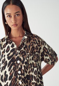 Kortärmad skjorta med knappar i krämfärg med leopardmönster i brunt och svart. Har en bröstficka och ett mjukt, texturerat tyg.