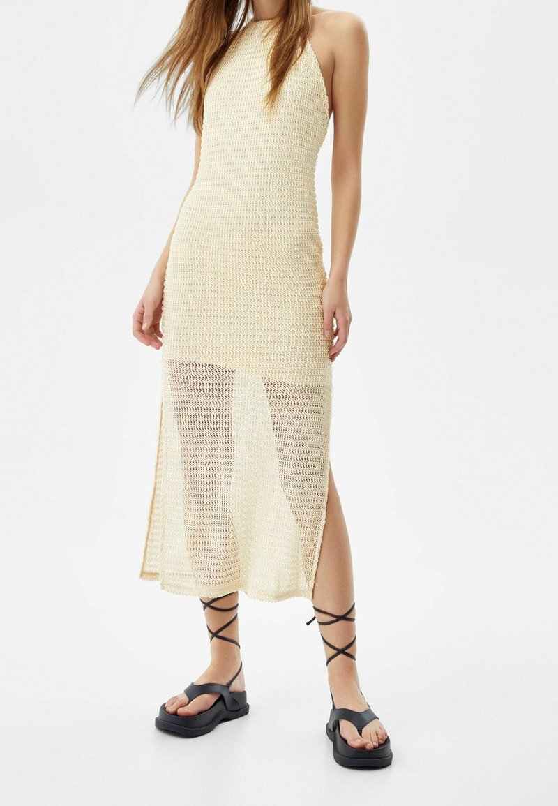 Pletené šaty s halter krkem v jemné béžové barvě s průhlednou spodní částí. Spárované s černými plochými sandály s křížovými řemínky.