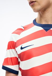 Személy piros-fehér csíkos Nike sportmezben, tengerészkék gallérral és ujjvégekkel, az orr alatt levágva.