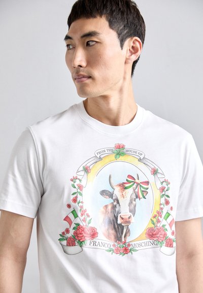 Balta medvilninė marškinėliai su spalvinga karvės grafika, apsupta gėlių ir juostelių, su tekstu "Iš Franco Moschino namų".