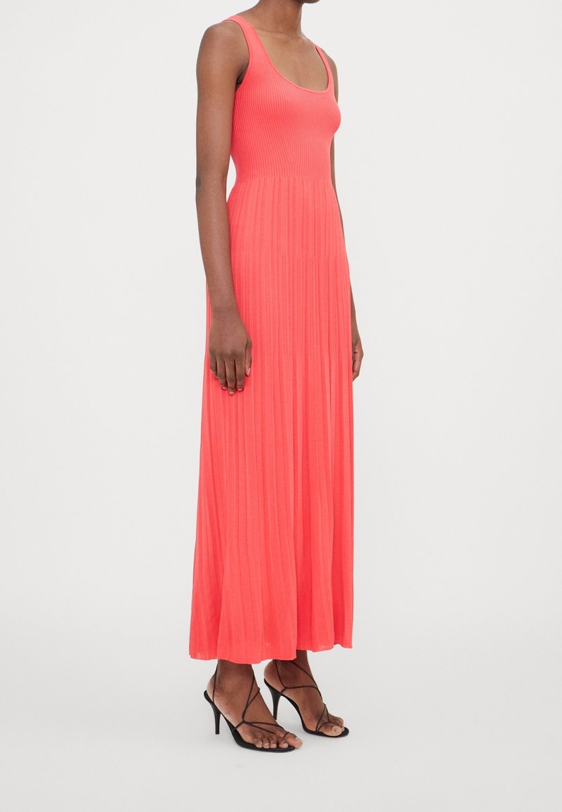 Femme portant une robe maxi corail plissée sans manches avec des sandales noires à talons hauts et fines brides, debout sur fond blanc.