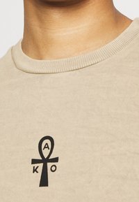 Beige bavlněné tričko s kulatým výstřihem. Vybaveno designem černého symbolu Ankh, do kterého jsou integrována písmena "A" a "O".