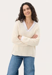 Cream-colored knit sweater with a V-neck, relaxed fit, and textured pattern, layered over a white collared shirt and paired with blue jeans.