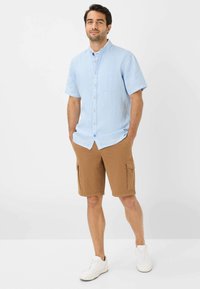 Camicia azzurra a maniche corte con bottoni e tasca sul petto, abbinata a pantaloni cargo marroni con tasche laterali e sneakers bianche.