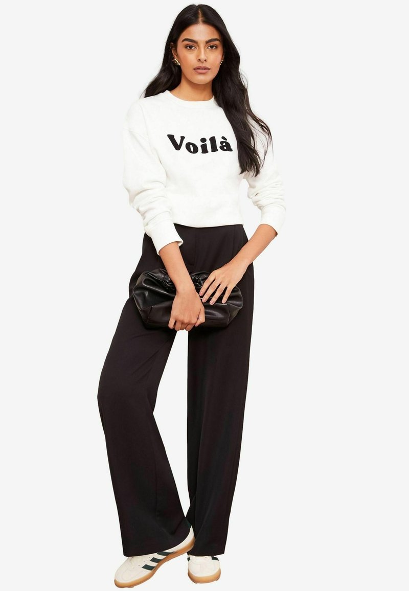 Felpa bianca con la scritta "Voilà" in nero, abbinata a pantaloni neri a vita alta e scarpe da ginnastica bianche con strisce nere. Pochette nera.
