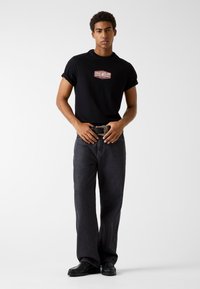 Zwart katoenen T-shirt met een rood-wit logo, gecombineerd met donker denim jeans en zwarte schoenen. Voorzien van opgerolde mouwen en een gestructureerde riem.