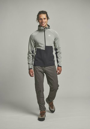 Mann lächelnd, trägt graue und schwarze Jacke mit Reißverschluss, graue Hose und beige Wanderschuhe, geht nach vorne vor schlichtem grauem Hintergrund.