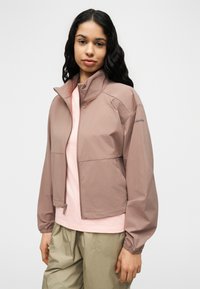 Jeune femme aux longs cheveux foncés portant une veste mauve à fermeture éclair, un haut rose clair et un pantalon vert olive sur un fond blanc uni.