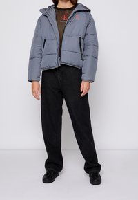 Szara kurtka ocieplana z kapturem, zapinana na zamek, z dwoma bocznymi kieszeniami. Noszona na brązowej koszulce i zestawiona z czarnymi szerokimi spodniami oraz czarnymi sneakersami.