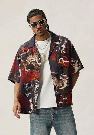 Uomo con capelli intrecciati che indossa occhiali da sole bianchi, camicia scura aperta con grandi stampe di gatti sopra una maglietta bianca e jeans blu, in piedi.