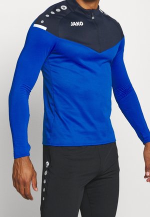 Mann trägt ein blau-schwarzes Sportoberteil mit langen Ärmeln und Frontreißverschluss sowie schwarze Sporthose mit runden Logos an den Seiten.