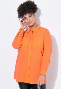 Camicia arancione brillante con un tessuto texture, tasche sul petto e maniche lunghe. Presenta un colletto classico e dettagli nei bottoni discreti.