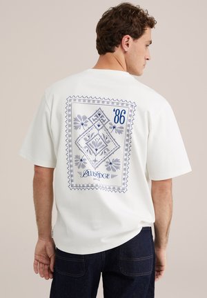 Mężczyzna o krótkich kręconych włosach, noszący białą, oversize'ową koszulkę z niebieskim geometrycznym wzorem przypominającym stempel i napisem "'86 Blue Ridge" na plecach.
