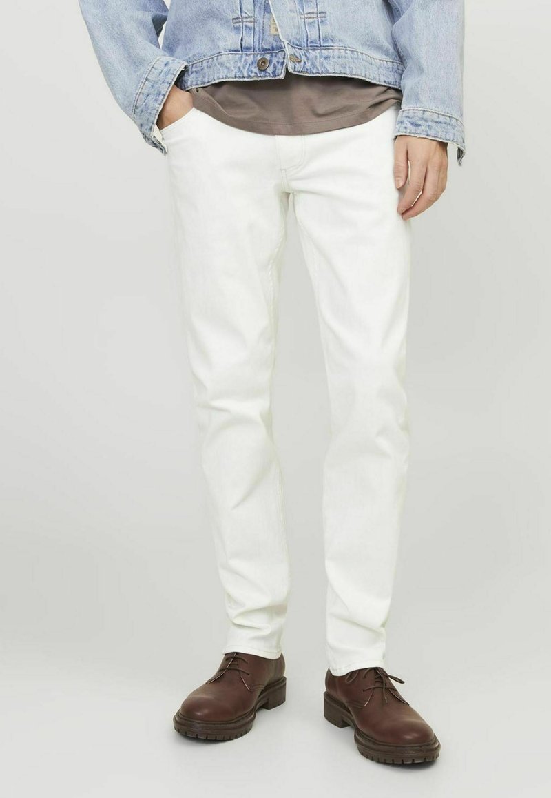 Jeans in denim bianco con vestibilità slim, abbinati a una camicia grigia e a una giacca di denim azzurro chiaro. Completati da scarpe stringate marroni.