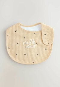 Bézs színű baba étkezőpárna bordázott textúrával, fekete pöttyös mintázattal és fehér hímzett "milk club" felirattal. Két patenttal rögzíthető.