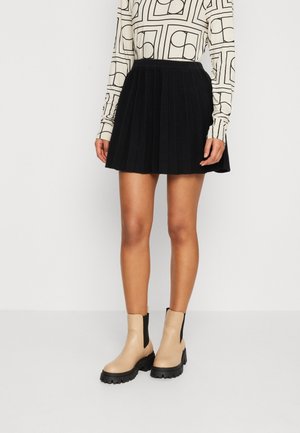 Femme portant une jupe plissée noire, des bottines beiges avec des côtés élastiques noirs, et un haut à manches longues blanc avec des motifs géométriques noirs.