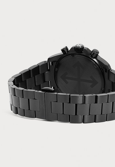 Montre-bracelet en métal noir avec un bracelet à maillons, montrant le boîtier arrière avec un logo gravé de flèches croisées et des boutons latéraux.
