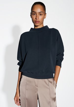 Vrouw met opgestoken haar, gekleed in een marineblauwe langemouwen top met een bijeengeknepen taille en beige satijnen wijde broek, staande tegen een witte achtergrond.