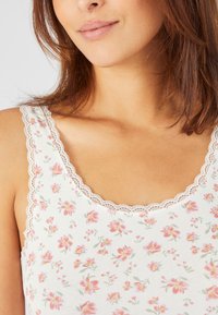 Débardeur fleuri avec un fond blanc, orné de fleurs roses et vertes. Il a un décolleté en dentelle festonnée et des bretelles fines. Texture du tissu douce.