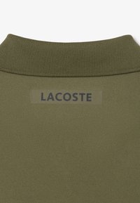 Olijfgroene polo met een gestructureerde, geribbelde kraag. Heeft een Lacoste logo patch in een contrasterende tan kleur op de achterkant van de halslijn.