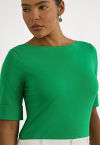 Πράσινο τοπ με κοντά μανίκια και φαρδύ λαιμόκοψη. Κατασκευασμένο από λείο ύφασμα με εφαρμοστή σιλουέτα. Διακριτικές μανσέτες και αδιάκοπος σχεδιασμός.