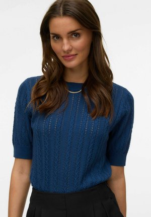 Femme aux longs cheveux bruns portant un pull court à manches courtes en tricot bleu et un pantalon noir taille haute, souriant devant un fond blanc uni.