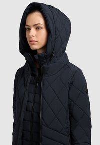 Gequilted navy pufferjack met een hoge kraag en grote capuchon. Voorzien van een rits aan de voorkant, zijzakken en een gestructureerde stofafwerking.