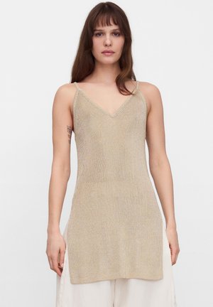 Junge Frau mit langem braunem Haar, die ein beiges ärmelloses Stricktop mit dünnen Trägern und V-Ausschnitt trägt, kombiniert mit lockeren weißen Hosen.