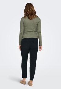 Olīvzaļš ribots ilgroku krekls ar ciešu piegriezumu, kombinēts ar melnām cieši pieguļošām biksēm. Redzams no aizmugures, izceļot gludo tekstūru un pieguļošo siluetu.