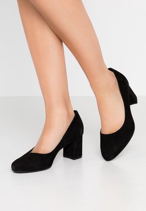 Jambes portant des escarpins en daim noir à talon bloc avec bout arrondi, debout sur un sol lisse gris clair sur un fond blanc uni.