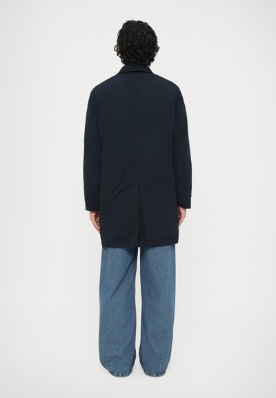 Mørkeblå lang frakke med snitkrave og fulde ærmer, parret med brede blå denimbukser. Glat tekstur, knælængde design.